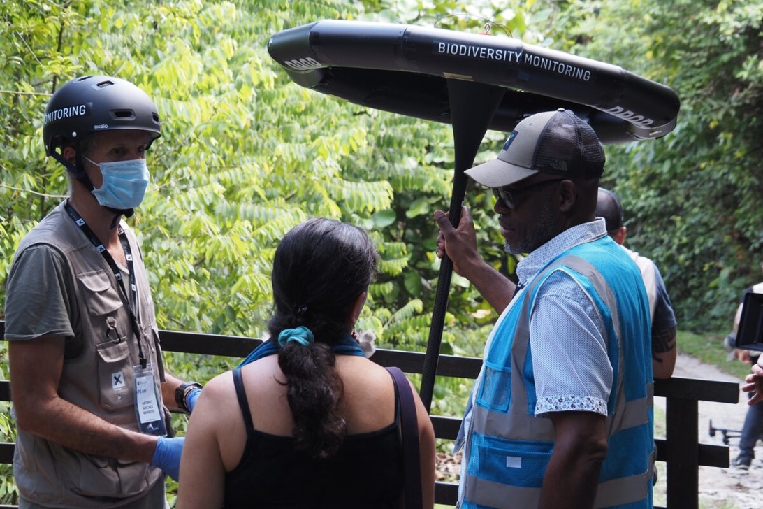 Field researchers in a tropical forest setting using professional biodiversity monitoring equipment labeled 'BIODIVERSITY MONITORING', with scientists examining the specialized technological device among lush green vegetation.